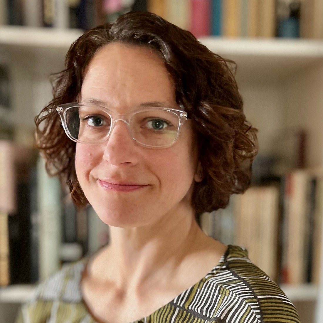 Aimee shapiro is a person with short brown hair that is slightly curly. she is wearing glasses with clear frames and has blue or light gree eyes.