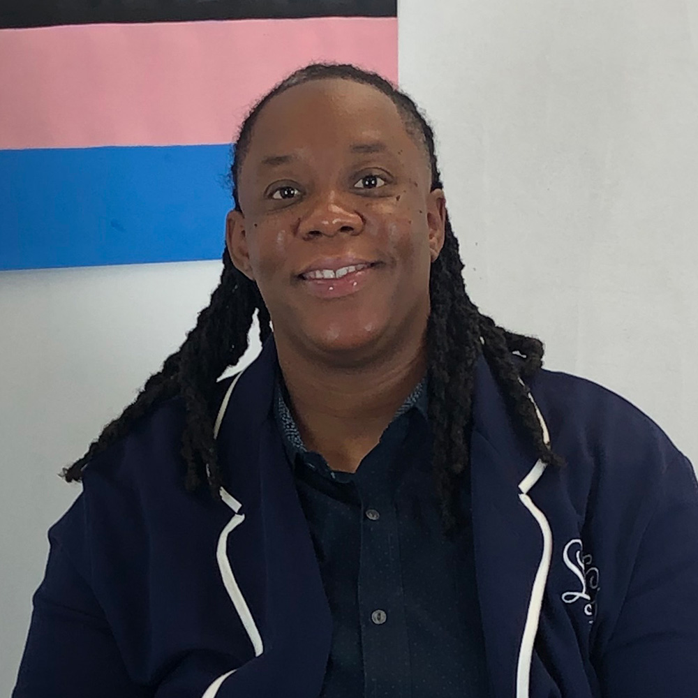 dana johnson is a person of color wearing a dark blue blazer with white trim. they have long thick braids.