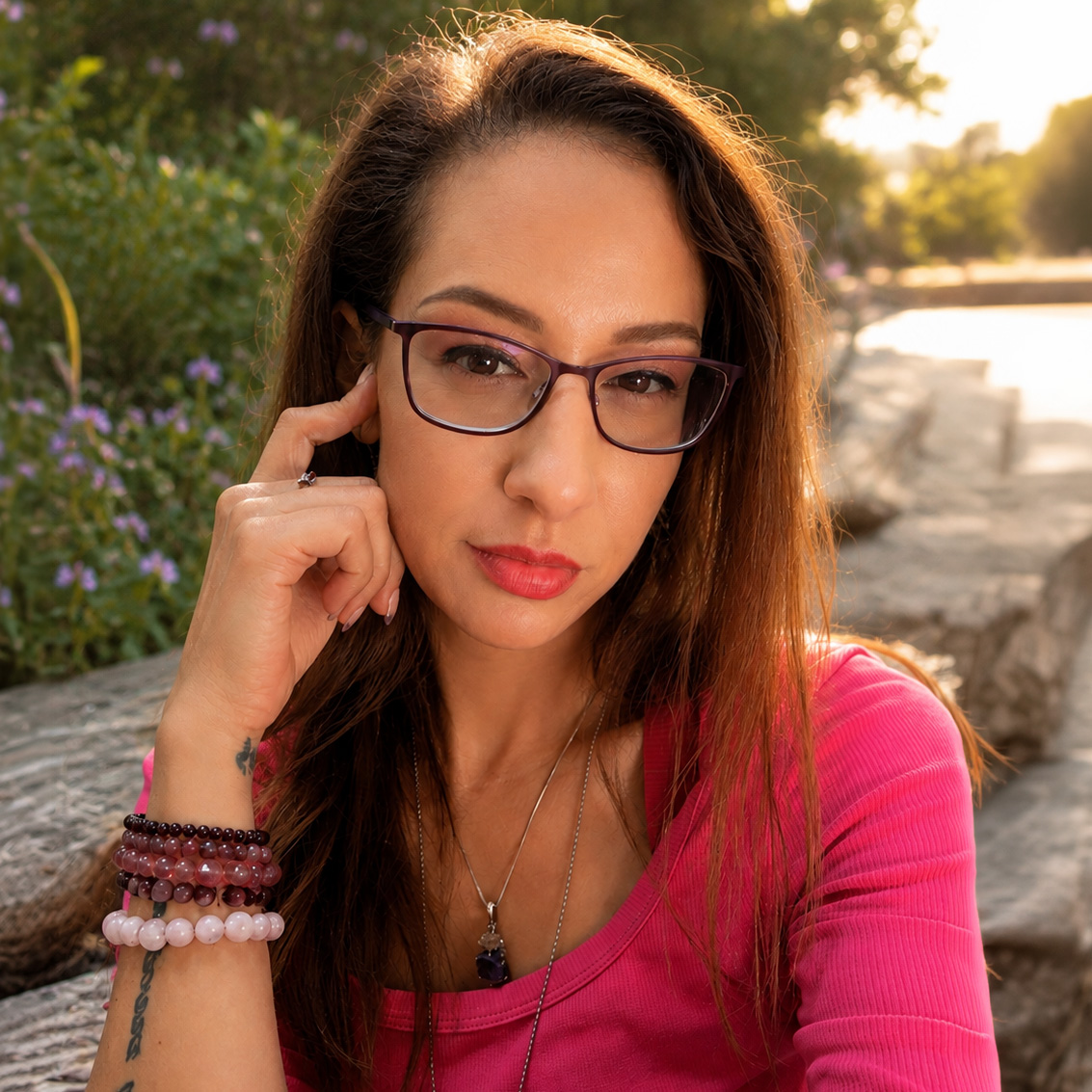 Loryn has shoulder-length hair brown hair and wears glasses. she is wearing a long-sleeved hot-pink top and is sitting near a stream