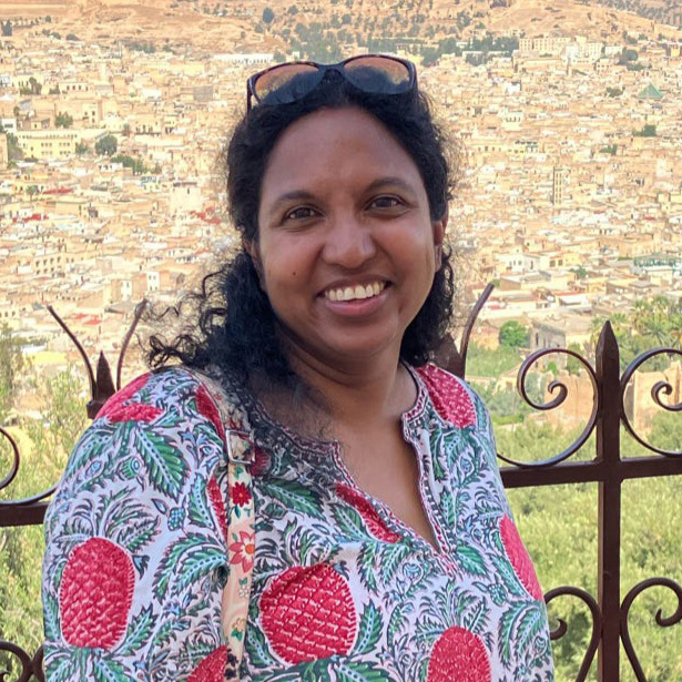 Nitara is a woman with shoulder length hair. she is wearing a pineapple printed cotton top and is posing in front of a scenic vista in a foreign country