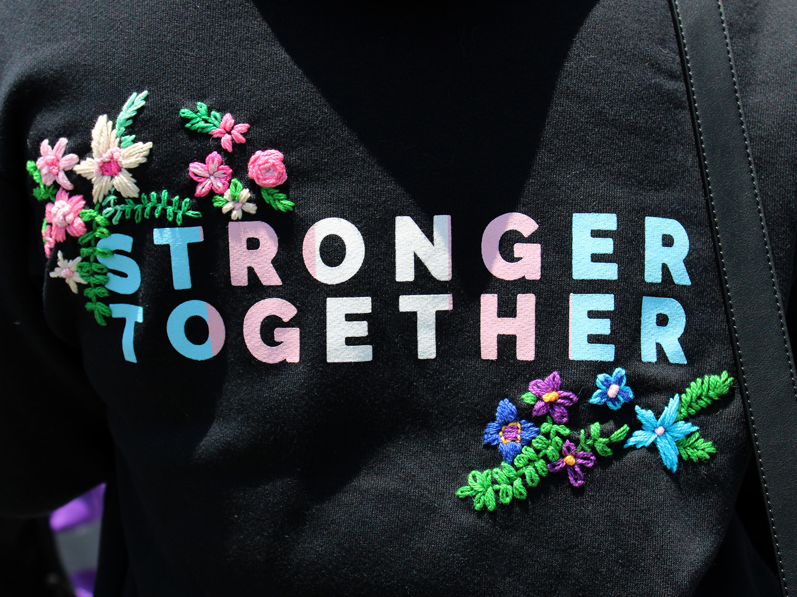 black coastpride sweatshirt that reads “stronger together” in trans flag color. there is flowery hand embroidery around the type
